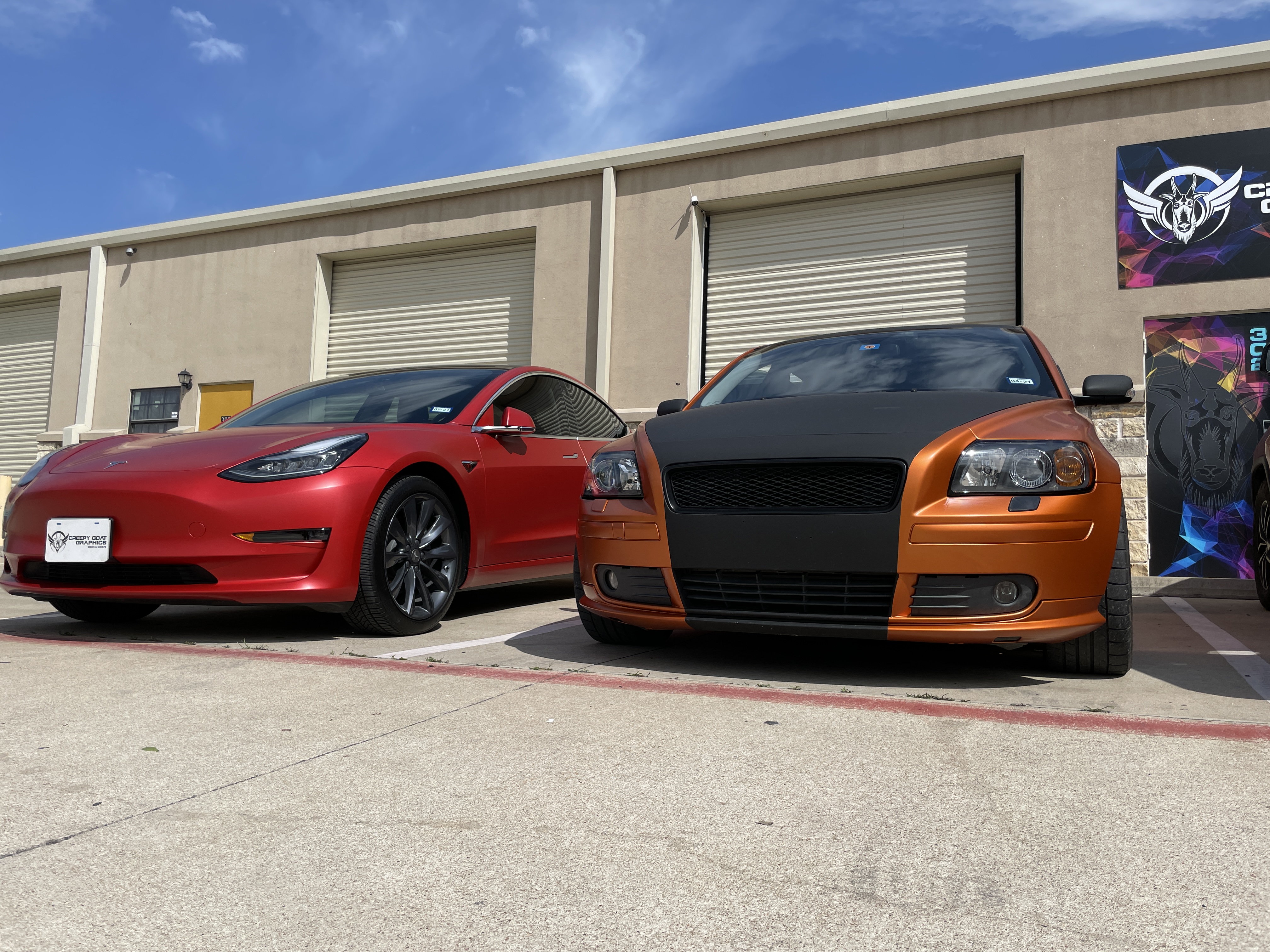 Wrapped gloss red Tesla parked next to gloss orang and black Volvo in from of Creepy Goat Graphics sign and wrap shop