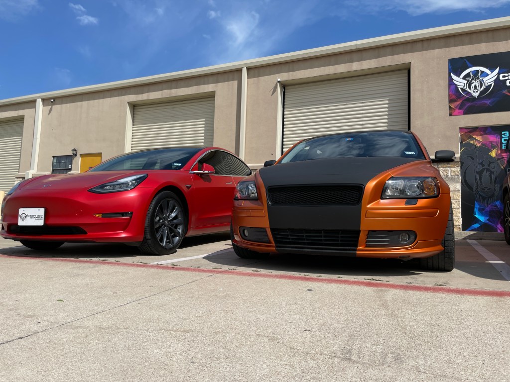 Wrapped gloss red Tesla parked next to gloss orang and black Volvo in from of Creepy Goat Graphics sign and wrap shop