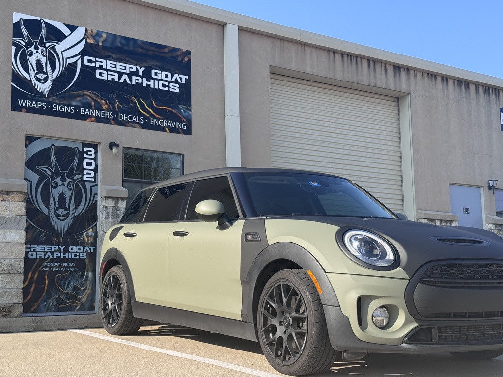 Custom matte khaki green Mini Cooper wrap parked outside Creepy Goat Graphics sign and wrap shop.